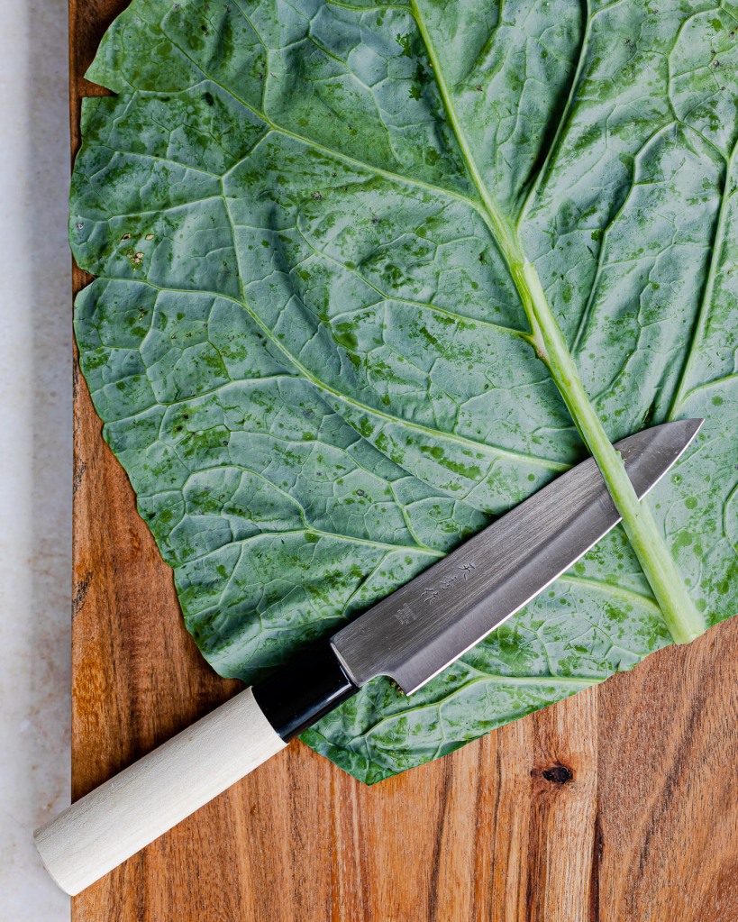 A collard leaf laying flat on a cutting board and a knife in the process of trimming away the excess thickness of the stem